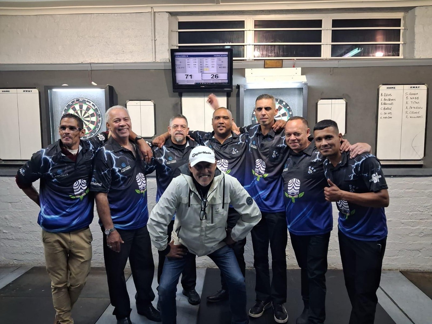 Eight Parklands players in club shirts in front of the dartboards and the match scoreboard